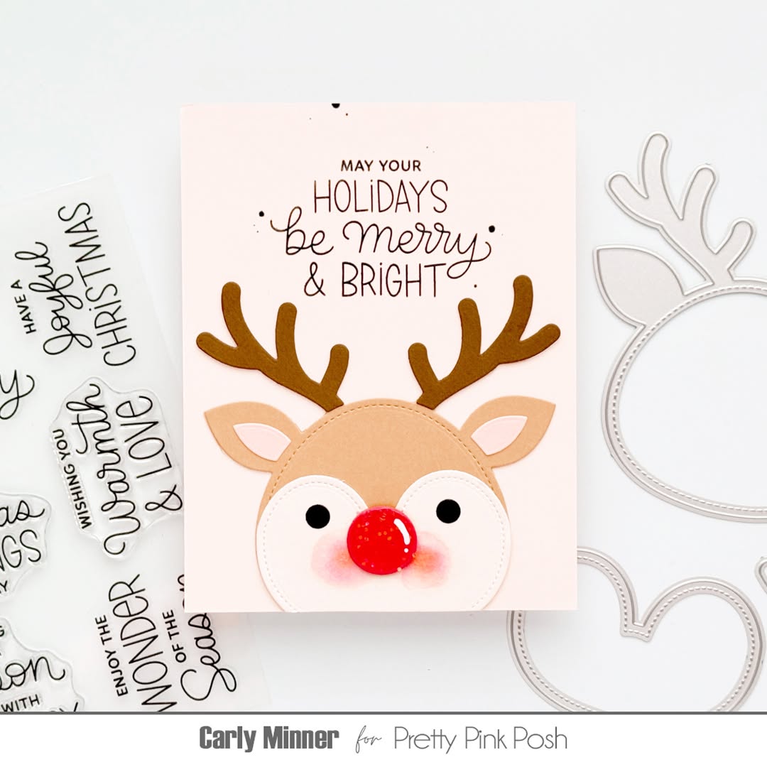 A handmade Christmas greeting card featuring a reindeer face with brown antlers, a red nose, and the text 'MAY YOUR HOLIDAYS Be Merry & BRIGHT' on a pale pink background. The card is styled on a white surface with crafting dies and text stamps visible around it.