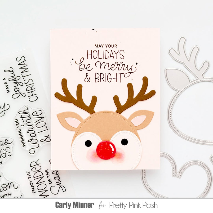 A handmade Christmas greeting card featuring a reindeer face with brown antlers, a red nose, and the text 'MAY YOUR HOLIDAYS Be Merry & BRIGHT' on a pale pink background. The card is styled on a white surface with crafting dies and text stamps visible around it.