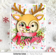 A handmade Christmas greeting card featuring a reindeer face with brown antlers, a pink and red scarf, and multicolored confetti inside the eyes. The card is displayed on a white textured background with festive holiday props, and the text 'Catherine Cohen for Pretty Pink Posh' appears at the bottom.