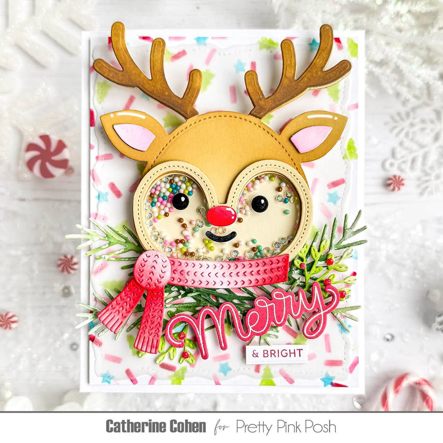 A handmade Christmas greeting card featuring a reindeer face with brown antlers, a pink and red scarf, and multicolored confetti inside the eyes. The card is displayed on a white textured background with festive holiday props, and the text 'Catherine Cohen for Pretty Pink Posh' appears at the bottom.