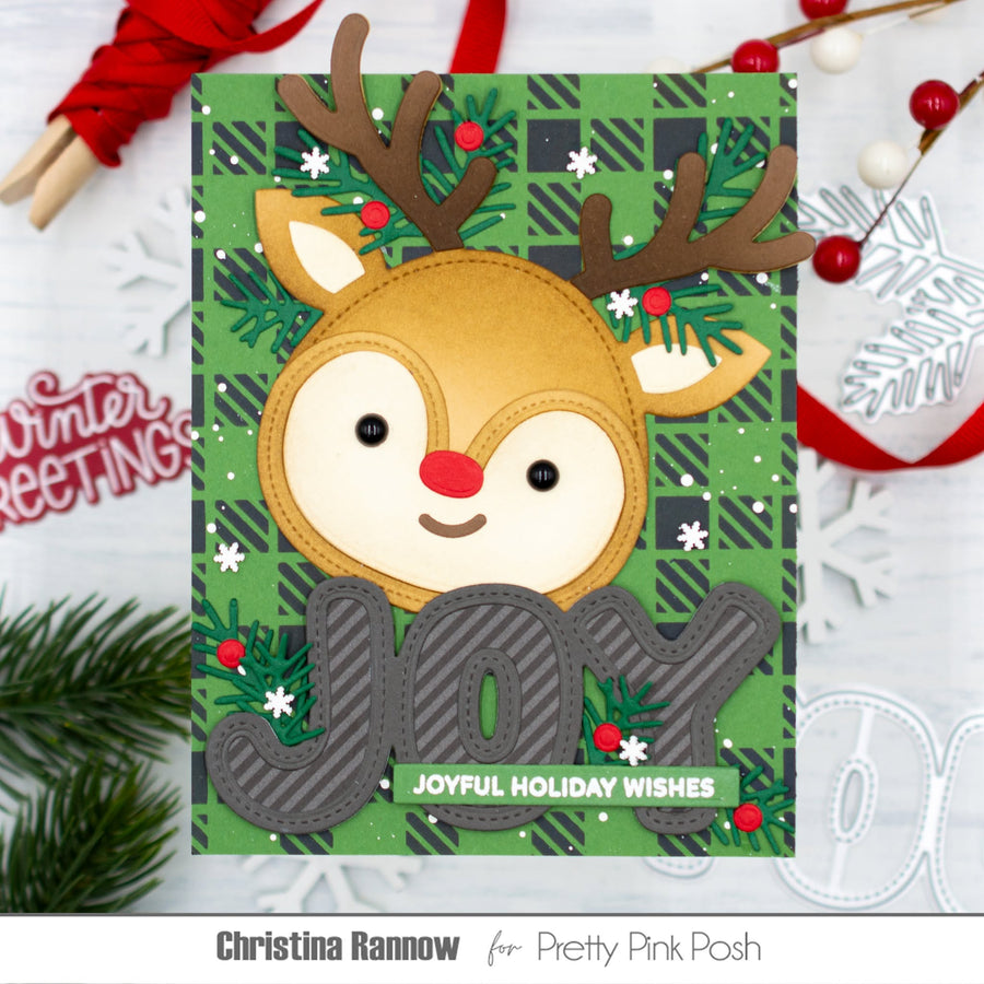 A handmade Christmas greeting card featuring a smiling reindeer face with brown antlers, a red nose, and a green plaid background. The card includes large gray layered letters spelling JOY and a small green banner reading 'JOYFUL HOLIDAY WISHES.