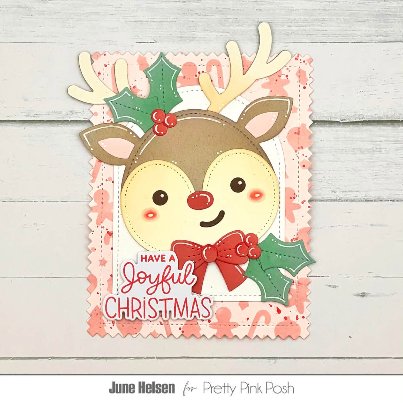 A handmade Christmas card featuring a cute reindeer face with antlers, green holly leaves, and red berries, with the text 'HAVE A Joyful CHRISTMAS' on a scalloped pink and white patterned card. The card is photographed on a light gray wood background, with 'June Helsen for Pretty Pink Posh' printed below.