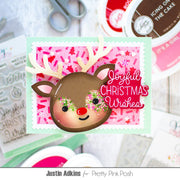 A handmade Christmas greeting card featuring a brown reindeer head with light antlers, a red nose, and glittery green and pink eyes on a pink patterned background. The card reads 'Joyful Christmas Wishes' in pink script.