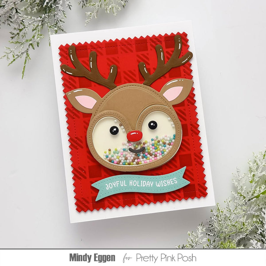 A handmade holiday greeting card featuring a reindeer face with brown antlers, a red nose, and colorful glitter in the eyes, set on a red patterned background with a teal banner reading 'Joyful Holiday Wishes'.