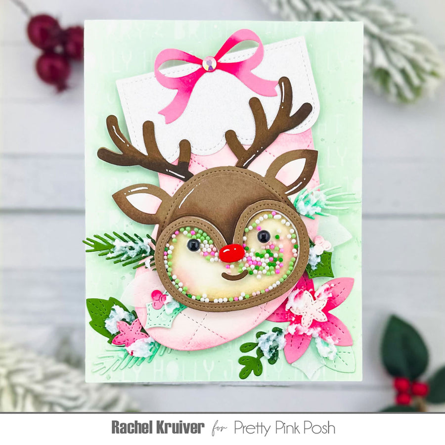 A handmade Christmas greeting card featuring a reindeer face with antlers, a pink bow, and festive floral details, photographed against a light wood background with holiday decorations.