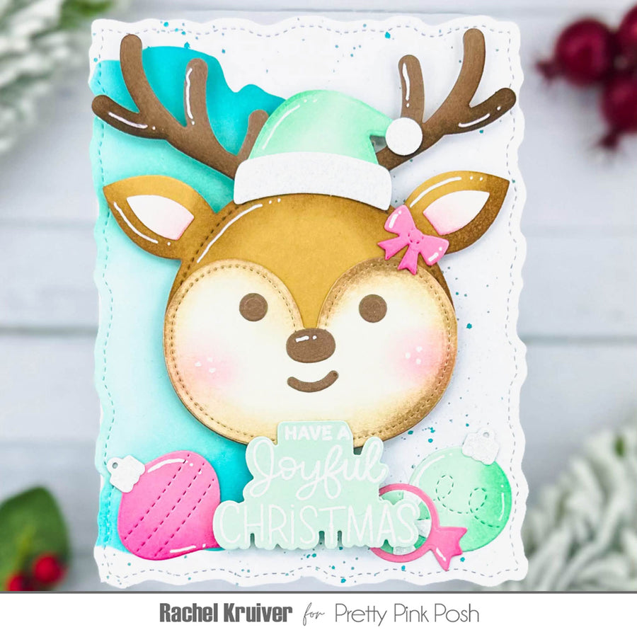 A handmade Christmas greeting card featuring a cute cartoon reindeer face with brown antlers, a mint green Santa hat, and a pink bow. The card has a scalloped white border, a light blue background, and includes the text 'HAVE A joyful CHRISTMAS' along with pink and mint ornaments.