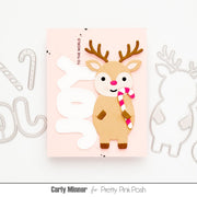 A handmade greeting card featuring a cute tan reindeer with brown antlers, a pink nose, and a pink-and-white striped candy cane, set against a light pink background with large white die-cut letters spelling 'JOY'. The card is displayed on a white surface with metal die-cutting templates partially visible on both sides.