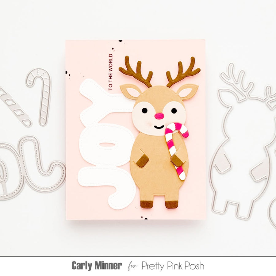 A handmade greeting card featuring a cute tan reindeer with brown antlers, a pink nose, and a pink-and-white striped candy cane, set against a light pink background with large white die-cut letters spelling 'JOY'. The card is displayed on a white surface with metal die-cutting templates partially visible on both sides.