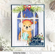 A handmade Christmas greeting card featuring a cute cartoon deer wearing white earmuffs and a red headband, holding a teal gift behind a window pane. The card is decorated with green pine branches, a red bow, and the text 'HAVE A Joyful CHRISTMAS' with holly accents.