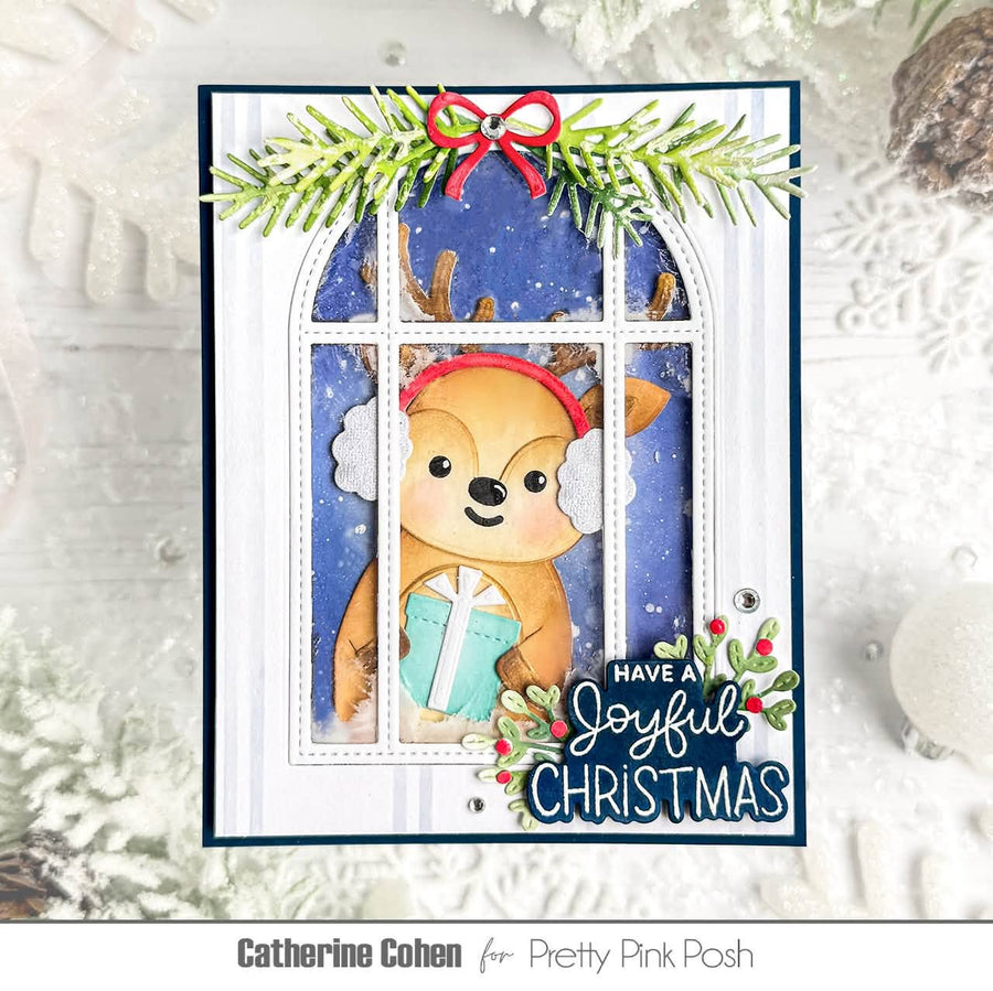 A handmade Christmas greeting card featuring a cute cartoon deer wearing white earmuffs and a red headband, holding a teal gift behind a window pane. The card is decorated with green pine branches, a red bow, and the text 'HAVE A Joyful CHRISTMAS' with holly accents.