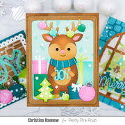 A handmade Christmas card featuring a cute brown reindeer with colorful antlers, a blue scarf, and the word 'JOY' on its chest, surrounded by presents, a green tree, and ornaments. The card is displayed on a white surface with other cards and ornaments in the background.
