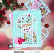 A handmade Christmas card featuring a reindeer behind a window with candy canes and red berries, styled with holiday ornaments and a card in the background. Text at the bottom reads 'Debbie Frank for Pretty Pink Posh'.