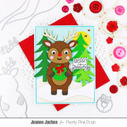 A handmade Christmas greeting card featuring a brown reindeer with antlers holding a green wreath, set against green trees and a yellow sun, with 'Merry Greetings' written on it. The card is styled on a white background with red envelopes and pink and red craft embellishments, with the text 'Jeanne Jachna for Pretty Pink Posh' at the bottom.