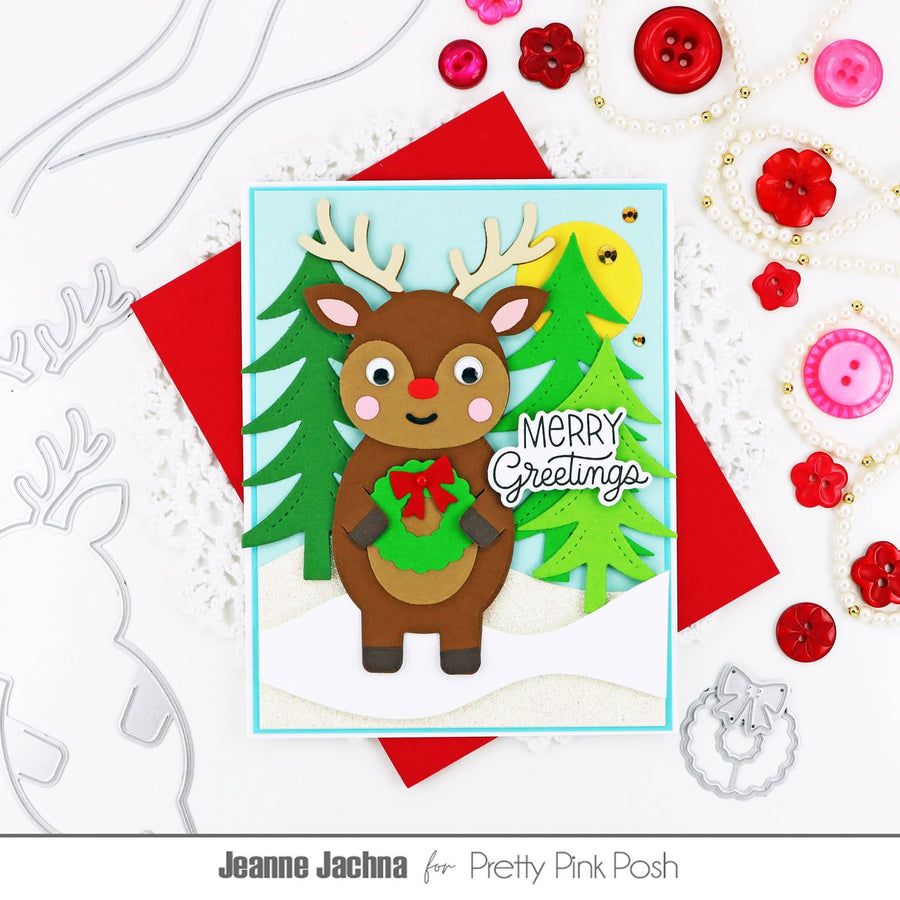 A handmade Christmas greeting card featuring a brown reindeer with antlers holding a green wreath, set against green trees and a yellow sun, with 'Merry Greetings' written on it. The card is styled on a white background with red envelopes and pink and red craft embellishments, with the text 'Jeanne Jachna for Pretty Pink Posh' at the bottom.