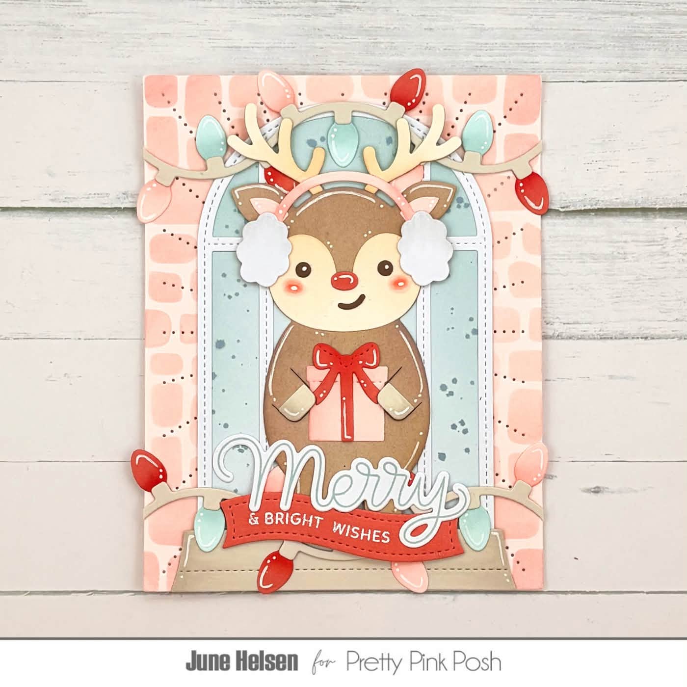 A handmade Christmas greeting card featuring a cute cartoon reindeer with white ear muffs, a red nose, and a red bow, set against a pastel pink and light blue patterned background with hanging red and light blue Christmas lights. The card is displayed on a light gray wood plank surface, with the text 'June Helsen for Pretty Pink Posh' printed below.