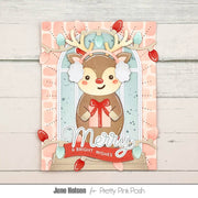 A handmade Christmas greeting card featuring a cute cartoon reindeer with white ear muffs, a red nose, and a red bow, set against a pastel pink and light blue patterned background with hanging red and light blue Christmas lights. The card is displayed on a light gray wood plank surface, with the text 'June Helsen for Pretty Pink Posh' printed below.