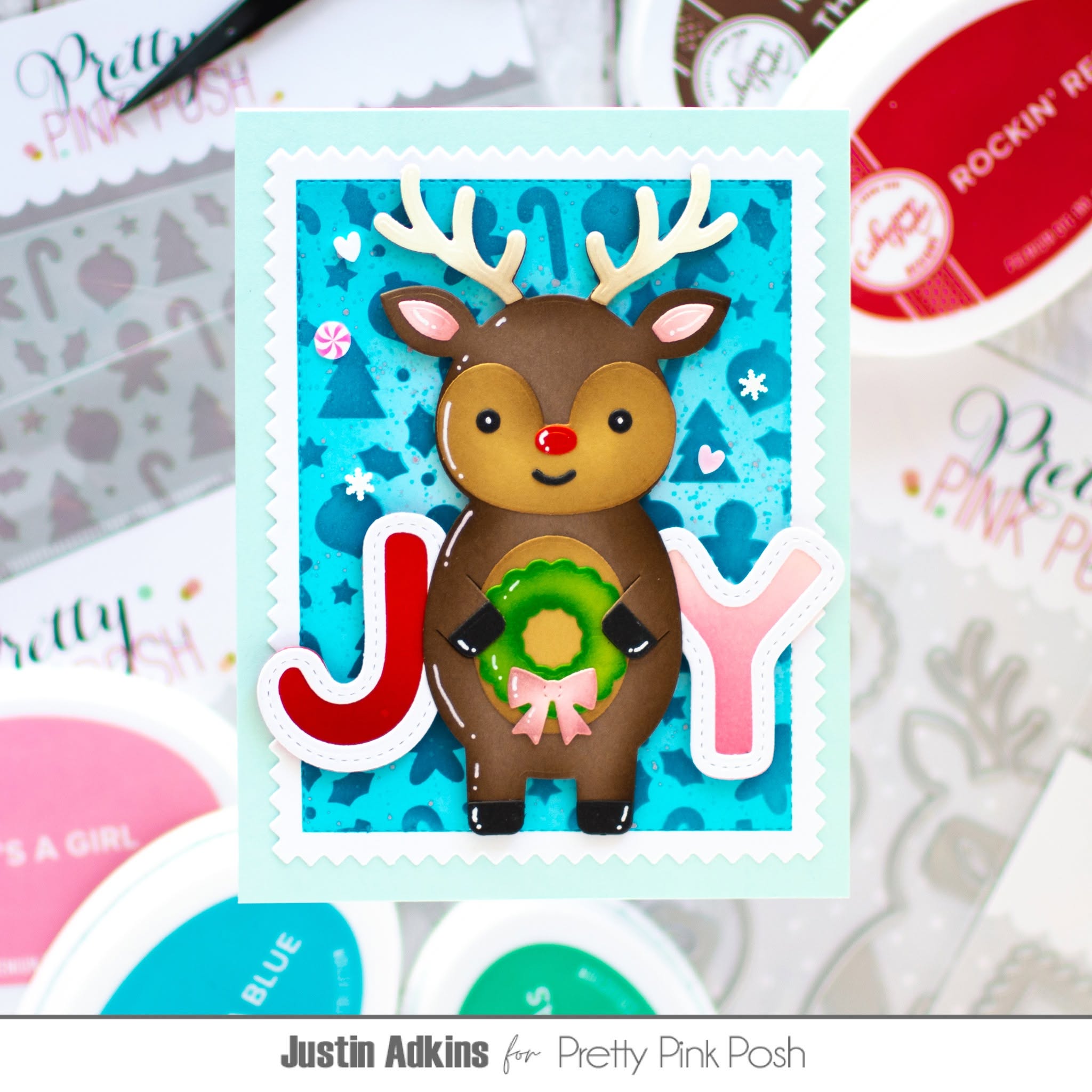 A handmade Christmas card featuring a brown reindeer with white antlers holding a green wreath, set against a blue patterned background with the letters J and Y in red and pink. The card is displayed on a styled flat-lay of craft supplies, including ink pads and stamps.