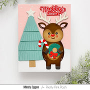 A handmade Christmas greeting card featuring a brown reindeer with antlers holding a candy cane and wreath, next to a teal Christmas tree, on a pink background with 'Merry & Bright' text. The card is displayed against a white wall with decorative greenery.