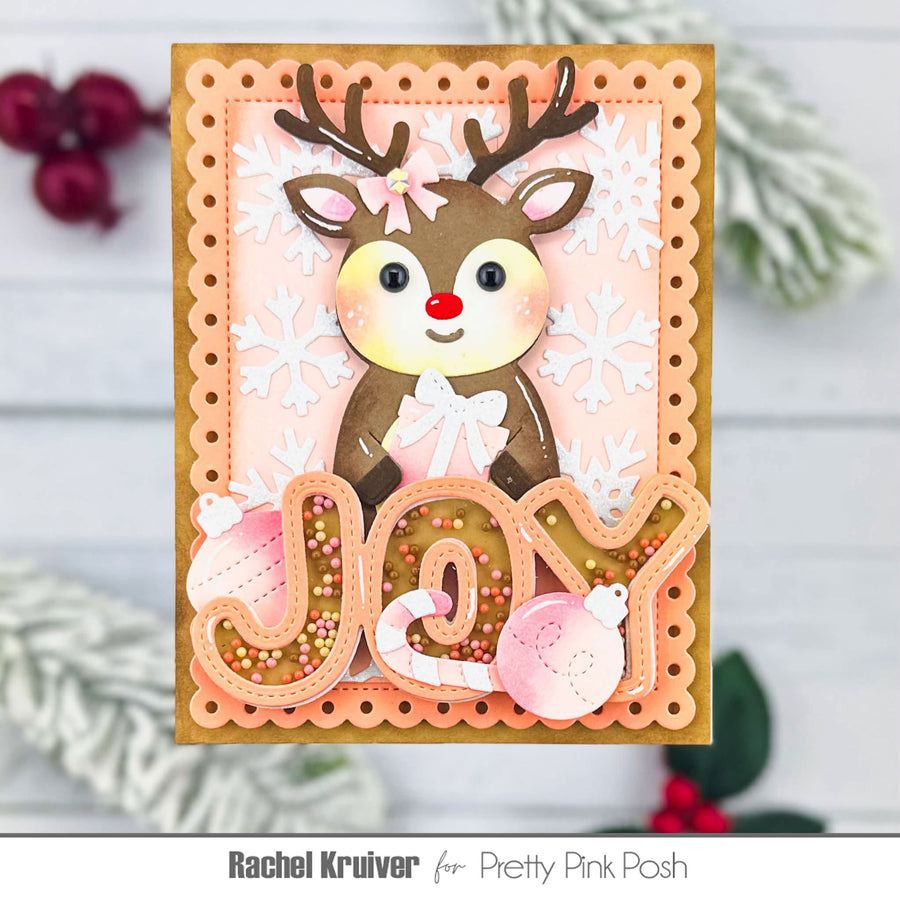 A handmade Christmas greeting card featuring a cute brown reindeer with antlers, a red nose, and a pink bow, layered over white die-cut snowflakes. The card has a scalloped peach border, a gold-toned inner frame, and 3D lettering spelling 'JOY' filled with pink ornaments and glitter.