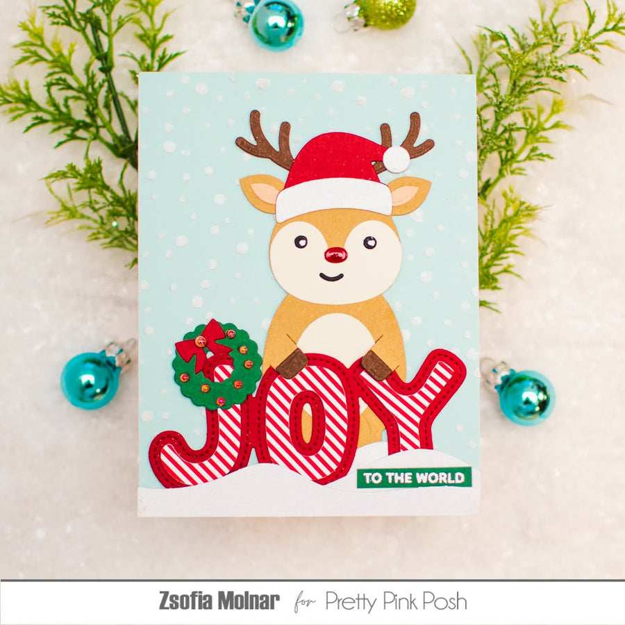 A handmade Christmas card featuring a cute cartoon reindeer in a Santa hat above the word 'JOY' in red-and-white striped letters, with a small green wreath and the phrase 'TO THE WORLD.' The card is styled on a white surface with greenery and blue ornaments around it.