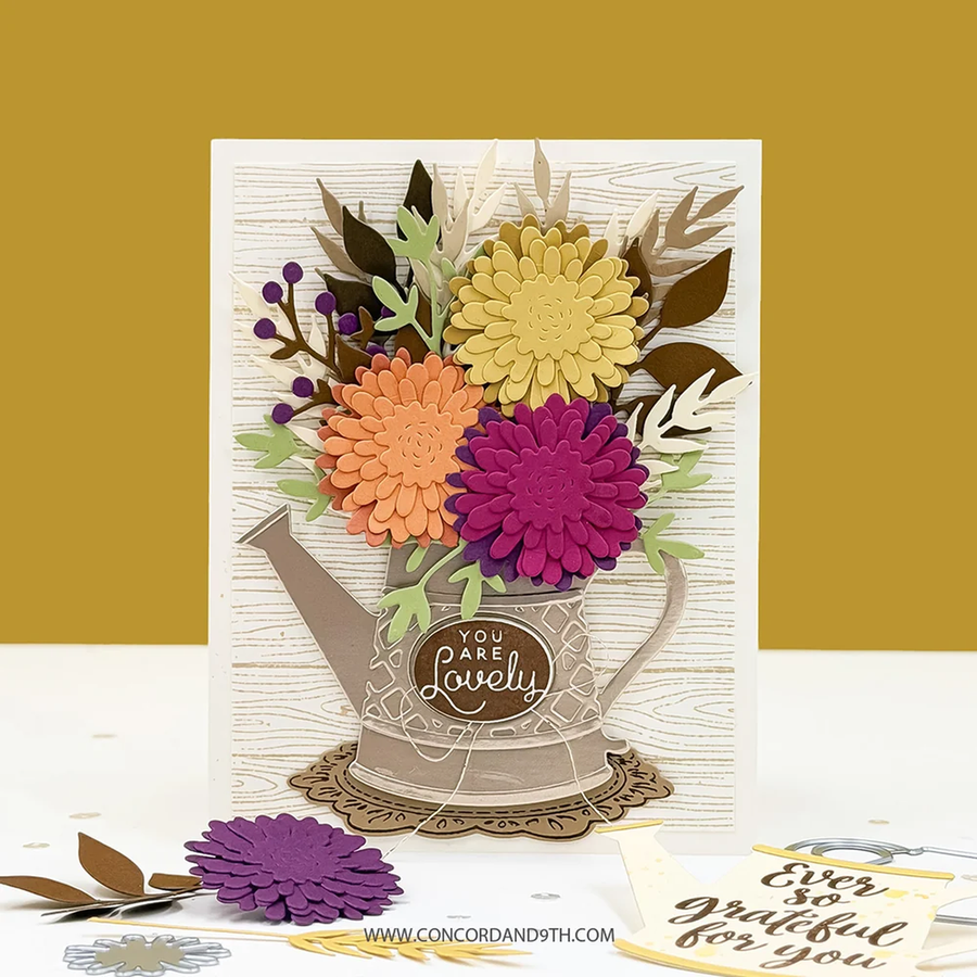 A handmade layered card featuring a watering can design with layered paper flowers in yellow, orange, and purple, set against a textured cream background. The card is displayed on a white surface with a mustard yellow background, with a loose purple flower and a small card in the foreground.
