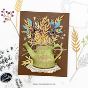A handmade layered greeting card featuring a green watering can shape with the words 'I am so grateful for you' in gold foil, filled with gold and blue leafy stems and small purple berries on a brown background. The card is styled on a white surface with craft stamps and die-cut elements around it.