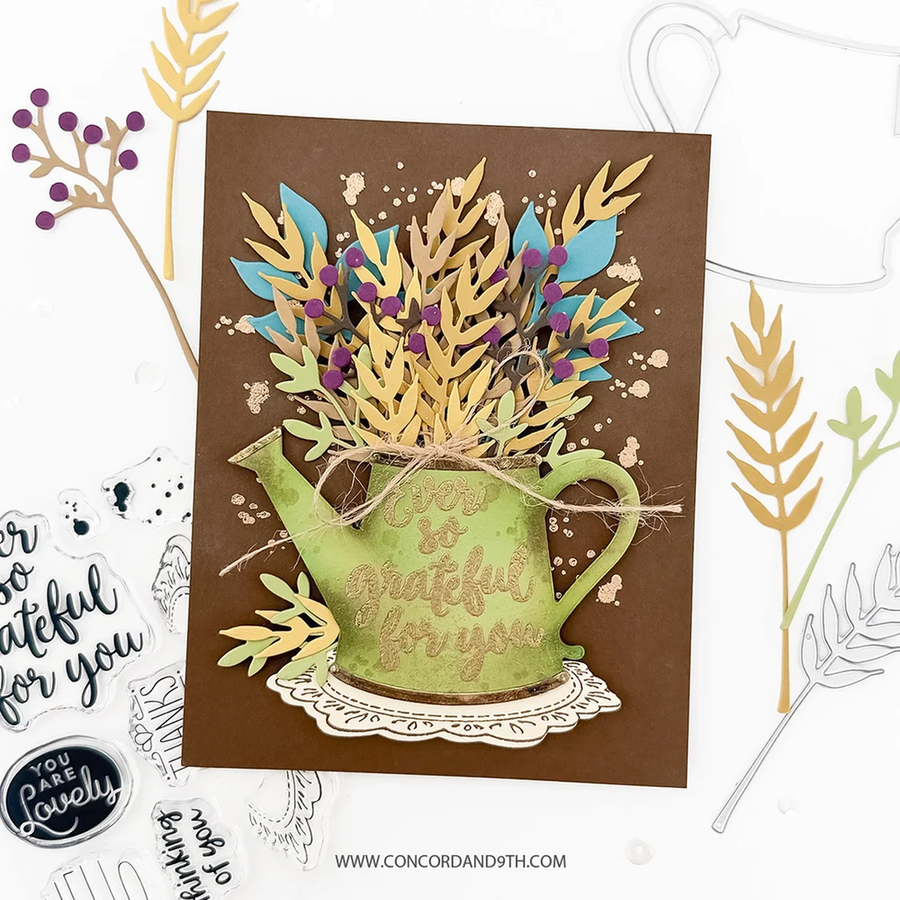 A handmade layered greeting card featuring a green watering can shape with the words 'I am so grateful for you' in gold foil, filled with gold and blue leafy stems and small purple berries on a brown background. The card is styled on a white surface with craft stamps and die-cut elements around it.