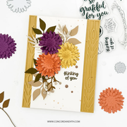 A handmade layered paper card with layered daisy flowers in purple, orange, and yellow, accented with green and beige leaves, set against a white background with crafting tools and words of gratitude.
