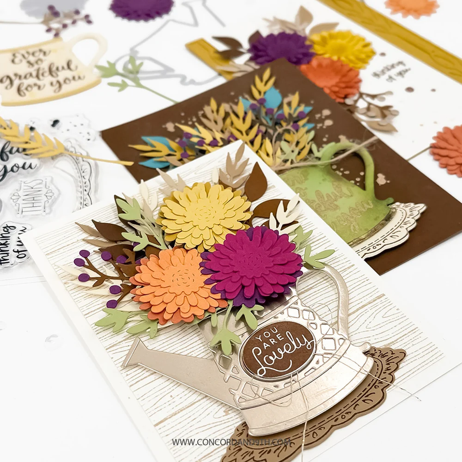 A close-up of a handmade layered paper card featuring a watering can design with layered paper flowers in yellow, orange, and magenta, green leaves, and a brown label reading 'You are loved.' Other cards and crafting elements are arranged around it on a white surface.