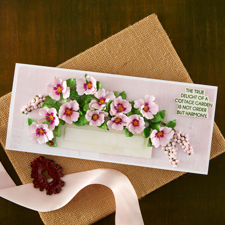 A rectangular handmade greeting card featuring pink flowers and green leaves, placed on a burlap surface with a pale pink ribbon and decorative seal nearby. The card includes printed text on the right side.