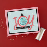 A handmade greeting card with a pink border and layered 'JOY' lettering, featuring a circular ornament design and a black banner that reads 'SENDING CHEER'. The card is displayed on a red background next to a spool of silver thread.