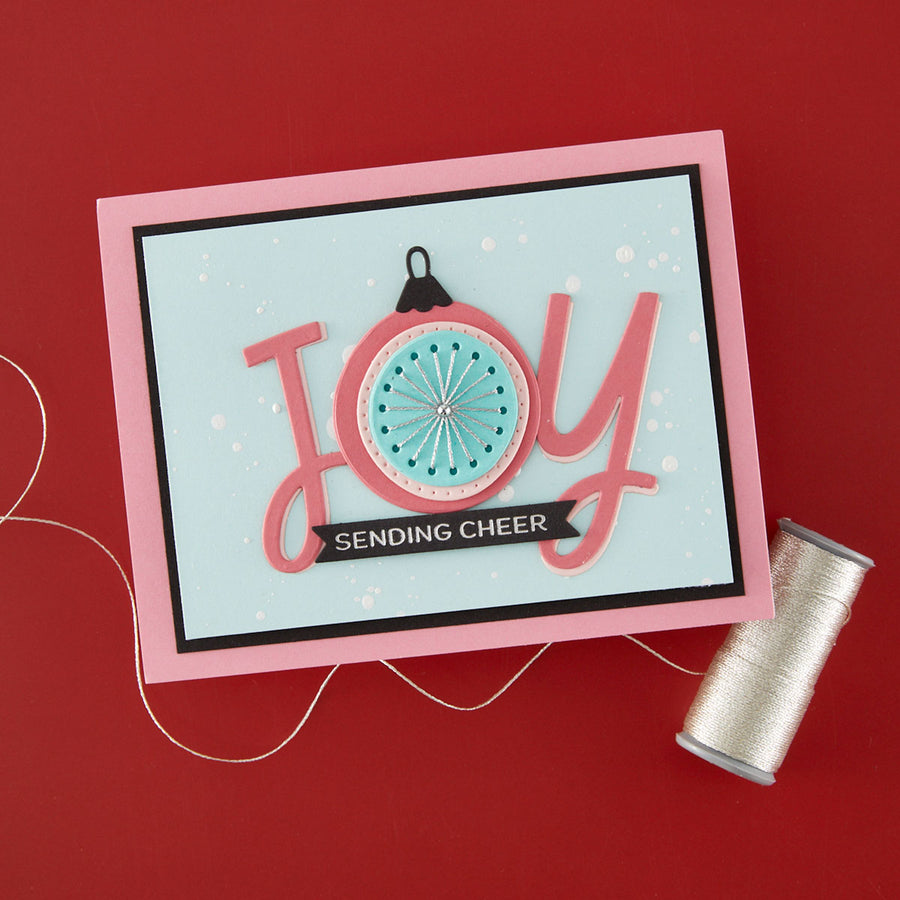 A handmade greeting card with a pink border and layered 'JOY' lettering, featuring a circular ornament design and a black banner that reads 'SENDING CHEER'. The card is displayed on a red background next to a spool of silver thread.