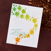 A handmade greeting card with layered die-cut maple leaves in green, yellow, and orange, along with white leaf cutouts and the script text 'Thinking OF YOU'. The card is photographed flat on a dark brown textured surface with dried brown leaves in the corner.