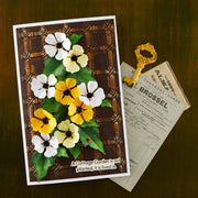 A handmade greeting card featuring 3D paper pansies in yellow, white, and pale yellow with green leaves, set against a brown lattice background. The card is styled on a dark wood surface next to a vintage gold key and an old newspaper page.