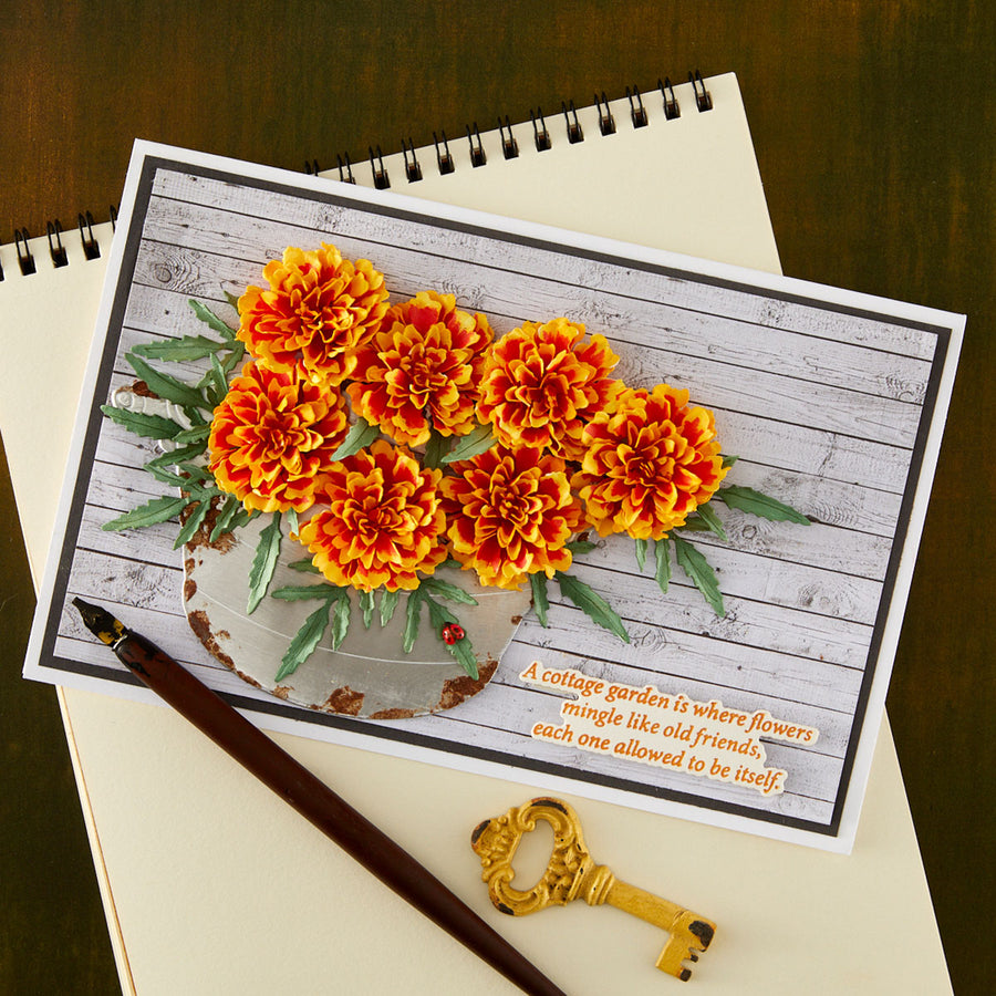 A handmade greeting card featuring a 3D pop-art design of orange and yellow marigold flowers in a rustic gray pot against a whitewashed wood background, with a quote printed in the lower right corner. The card is styled on a cream-colored spiral notebook with a gold key and a brown pen on a dark wooden table.