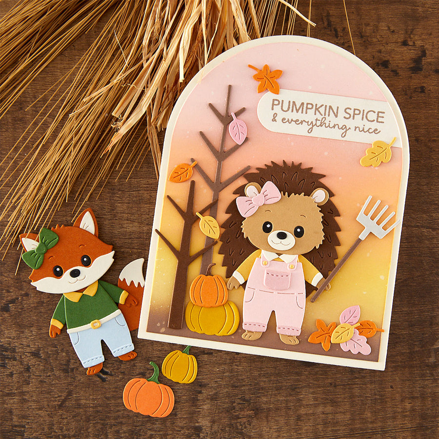 A handmade layered paper card featuring a hedgehog in pink overalls and a bow, standing beside a tree with autumn leaves and pumpkins, with the text 'PUMPKIN SPICE & everything nice.' A small fox figure in a green shirt and blue pants sits beside the card on a rustic wood surface with dried grass and loose paper cutouts.