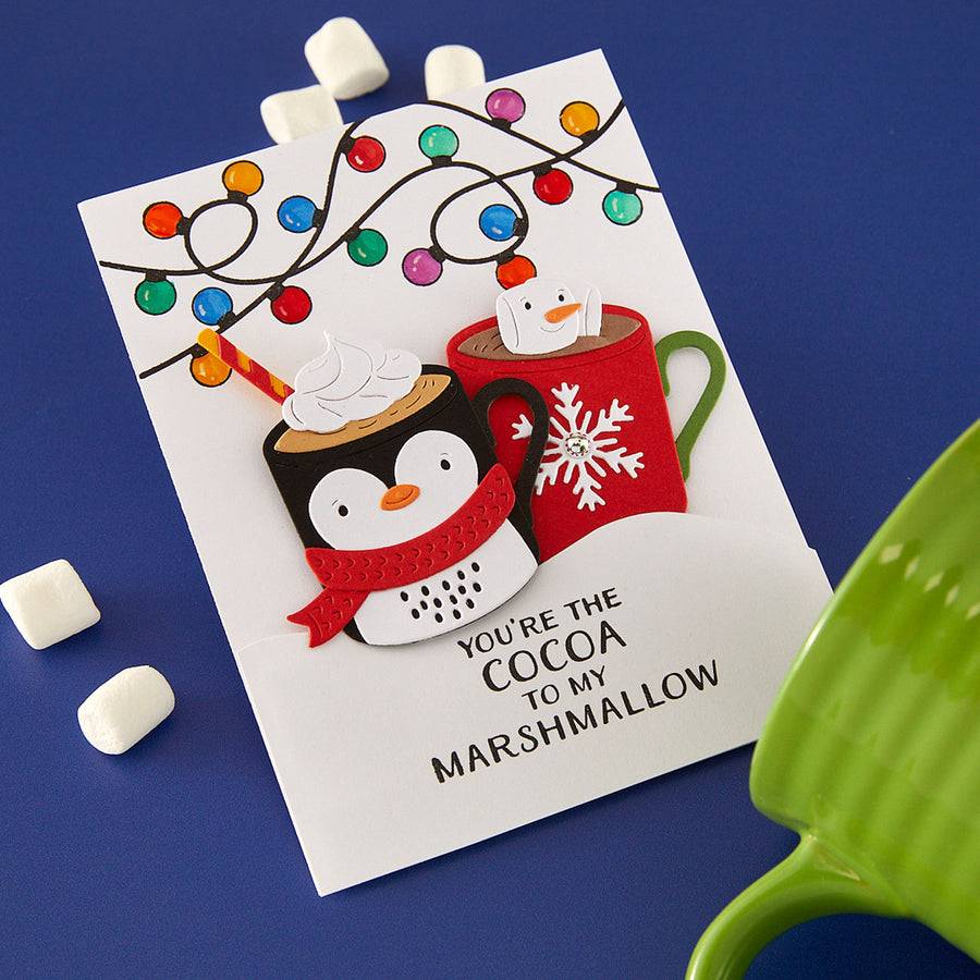 A handmade layered paper card featuring penguin cocoa cups under colorful string lights, with the text 'YOU'RE THE COCOA TO MY MARSHMALLOW', displayed on a blue background with marshmallows and a green mug prop. Spellbinders Mug Friends Etched Dies
