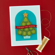 A handmade Christmas greeting card featuring a green layered tree design with a red banner reading 'CHERISH THE SEASON' on a light blue arched background. The card is photographed on a red surface with gold thread, scissors, and a spool of thread nearby.