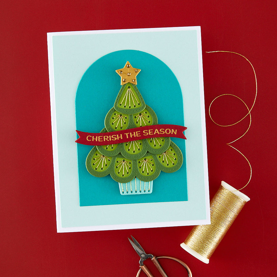 A handmade Christmas greeting card featuring a green layered tree design with a red banner reading 'CHERISH THE SEASON' on a light blue arched background. The card is photographed on a red surface with gold thread, scissors, and a spool of thread nearby.