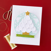 A handmade Christmas card featuring a layered die-cut white tree with a gold star and banner, set on a white embossed background. The card is styled on a red surface with gold scissors and a spool of gold thread nearby.