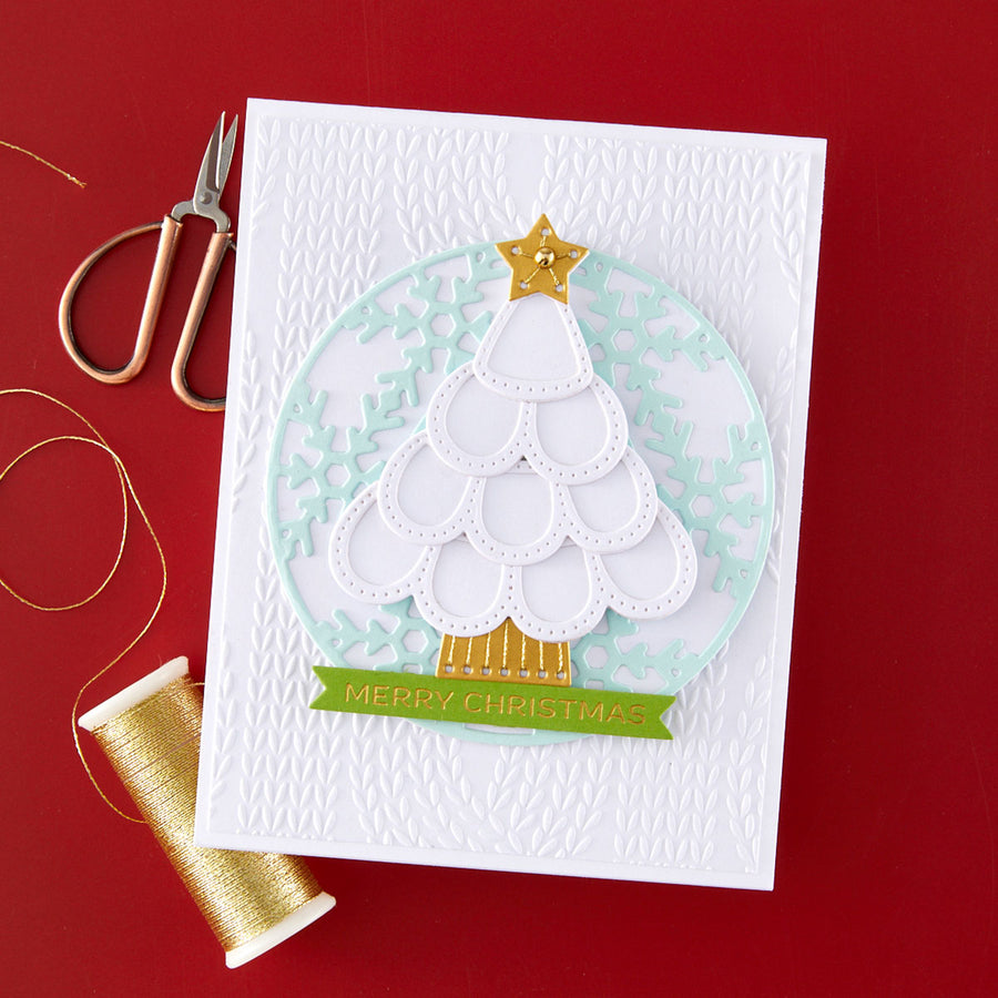 A handmade Christmas card featuring a layered die-cut white tree with a gold star and banner, set on a white embossed background. The card is styled on a red surface with gold scissors and a spool of gold thread nearby.