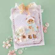 A decorative handmade card featuring a teddy bear dressed in a light blue winter outfit, surrounded by layered white and pink textured paper, embossed details, faux flowers, and a cream ribbon bow, set against a pale mint green background.