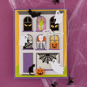 A layered Halloween-themed handmade greeting card featuring a haunted house design with windows showing ghostly eyes, a skeleton, a pumpkin, a spiderweb, a black cat, and floating white ghosts. The card is displayed on a purple background with spiderweb texture and black spider cutouts.
