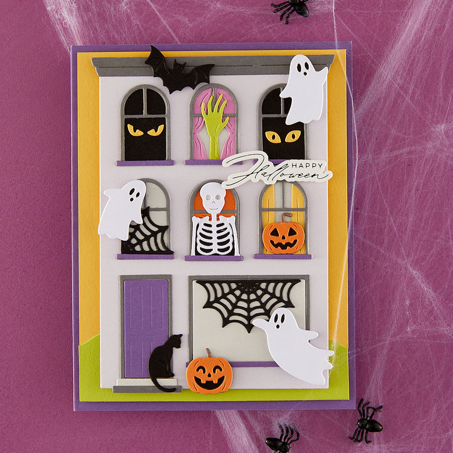 A layered Halloween-themed handmade greeting card featuring a haunted house design with windows showing ghostly eyes, a skeleton, a pumpkin, a spiderweb, a black cat, and floating white ghosts. The card is displayed on a purple background with spiderweb texture and black spider cutouts.