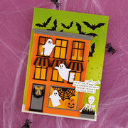 A handmade layered paper greeting card with a Halloween theme, featuring an orange haunted house, white ghosts, black bats, a spiderweb window, and a skeleton figure on a green-to-yellow gradient background, displayed on a textured pink surface.