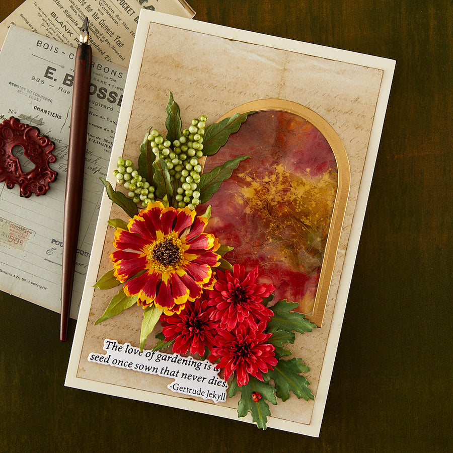 A handmade layered greeting card featuring red and yellow flowers, green leaves, and a gold-toned arched window overlay with a quote by Gertrude E. Bell, styled on a dark wood background next to a pen and vintage paper.