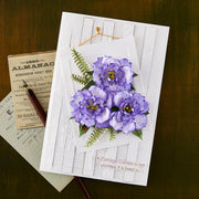 A handmade greeting card with a white wood-plank style base, featuring three purple flowers with green leaves, placed on top of vintage book pages and a pen on a dark olive background.
