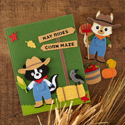 A rustic autumn-themed felt craft or book cover featuring a green background with a wooden sign reading 'HAY RIDES' and 'CORN MAZE,' plus felt animals including a skunk, fox, crow, corn, pumpkins, and wheat. The item is photographed on a wooden surface with styled seasonal props.