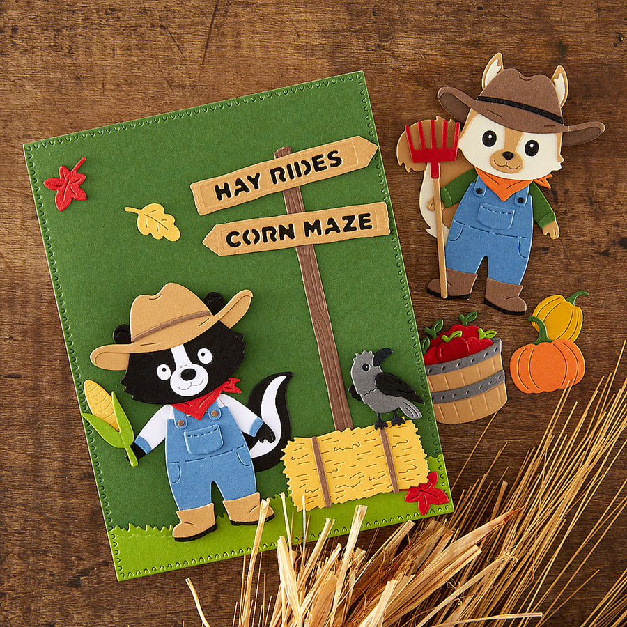 A rustic autumn-themed felt craft or book cover featuring a green background with a wooden sign reading 'HAY RIDES' and 'CORN MAZE,' plus felt animals including a skunk, fox, crow, corn, pumpkins, and wheat. The item is photographed on a wooden surface with styled seasonal props.