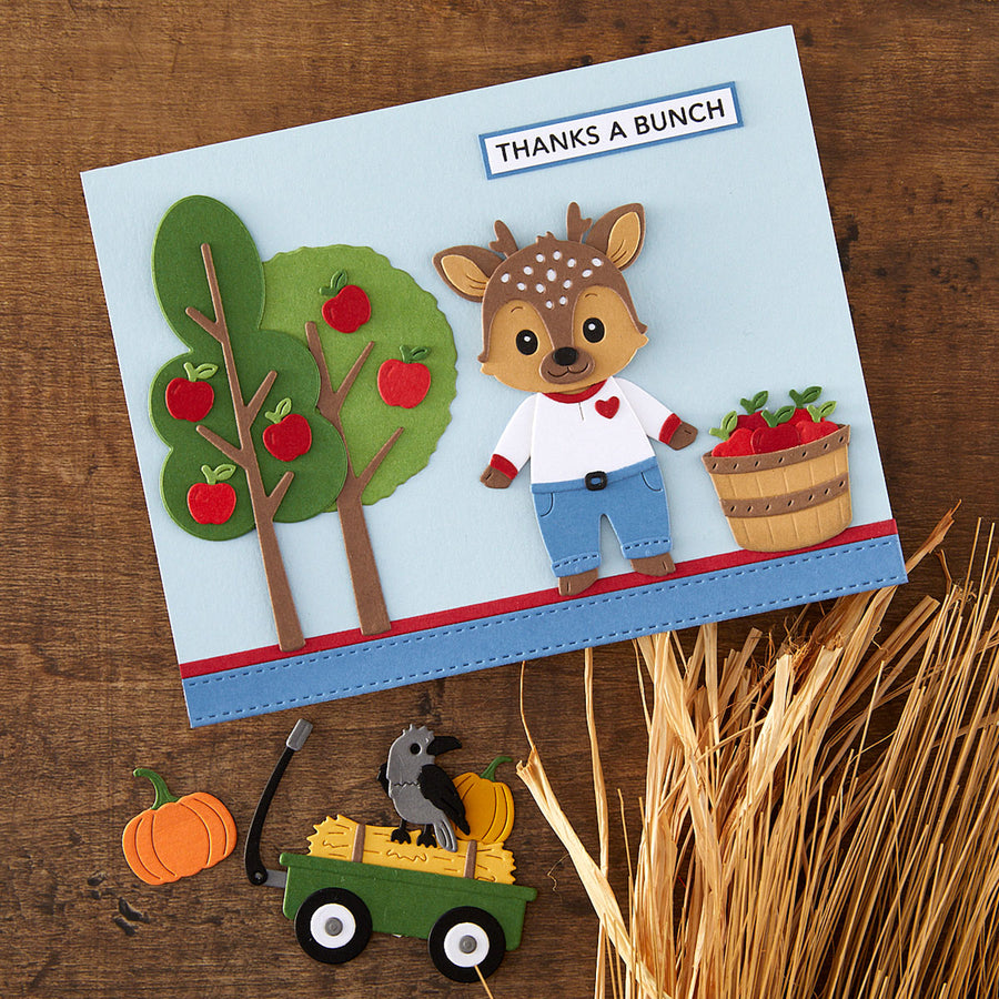 A handmade autumn-themed greeting card featuring a cute deer character, a tree with red apples, and a basket of strawberries, displayed on a wooden surface with seasonal paper cutouts.
